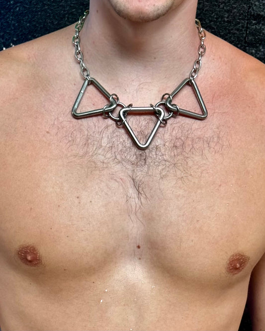 A person wearing a bold, geometric stainless steel necklace featuring three large, triangular links connected by chain rings. The necklace sits snugly on the person’s chest, showcasing the metallic design against bare skin.