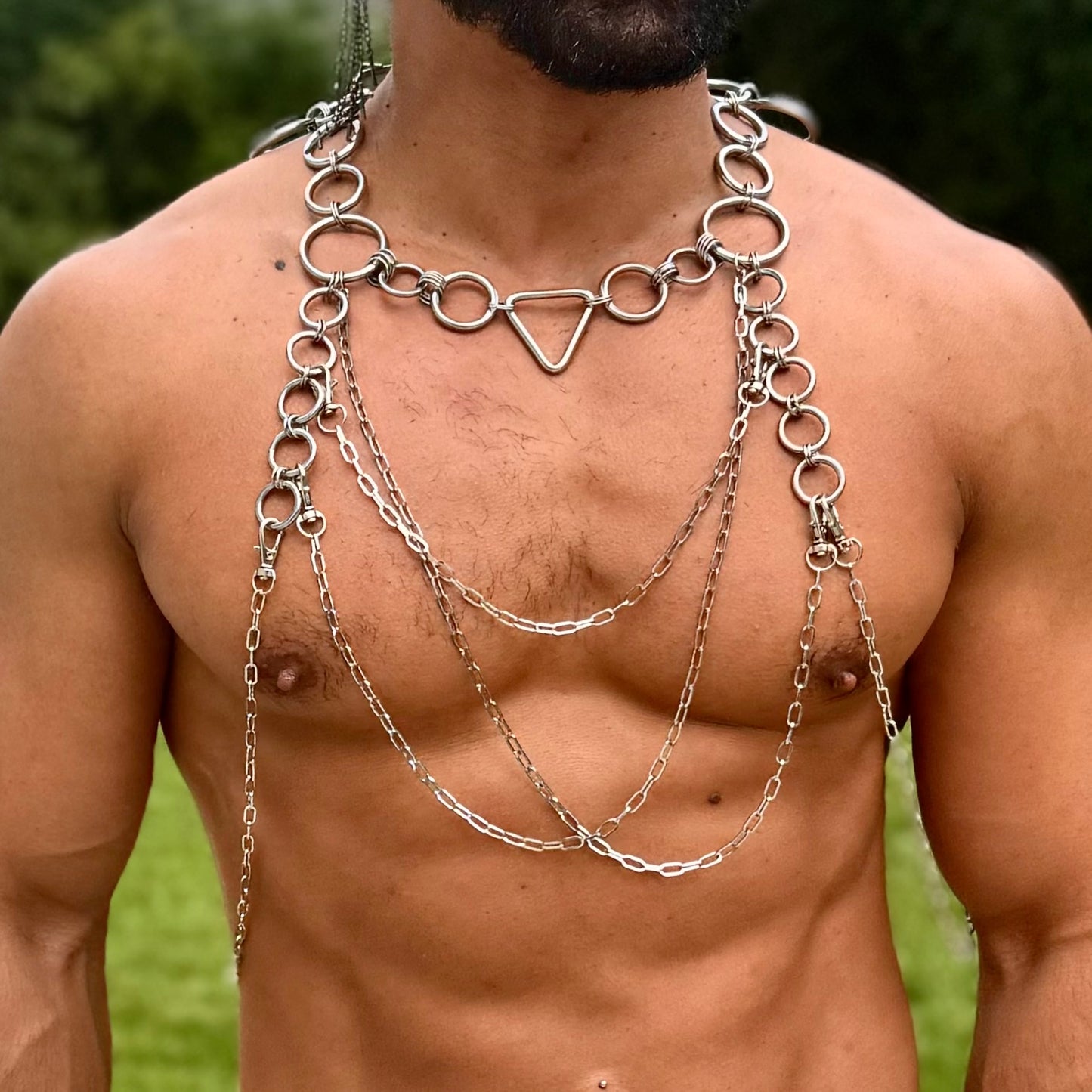 A person wearing an intricate body harness made of stainless steel rings and chains. The harness features a central triangle link at the neck, with multiple layers of chains draping across the chest and shoulders, creating a striking and artistic look. The design is bold, with circular rings connecting the chains and providing structure to the piece.