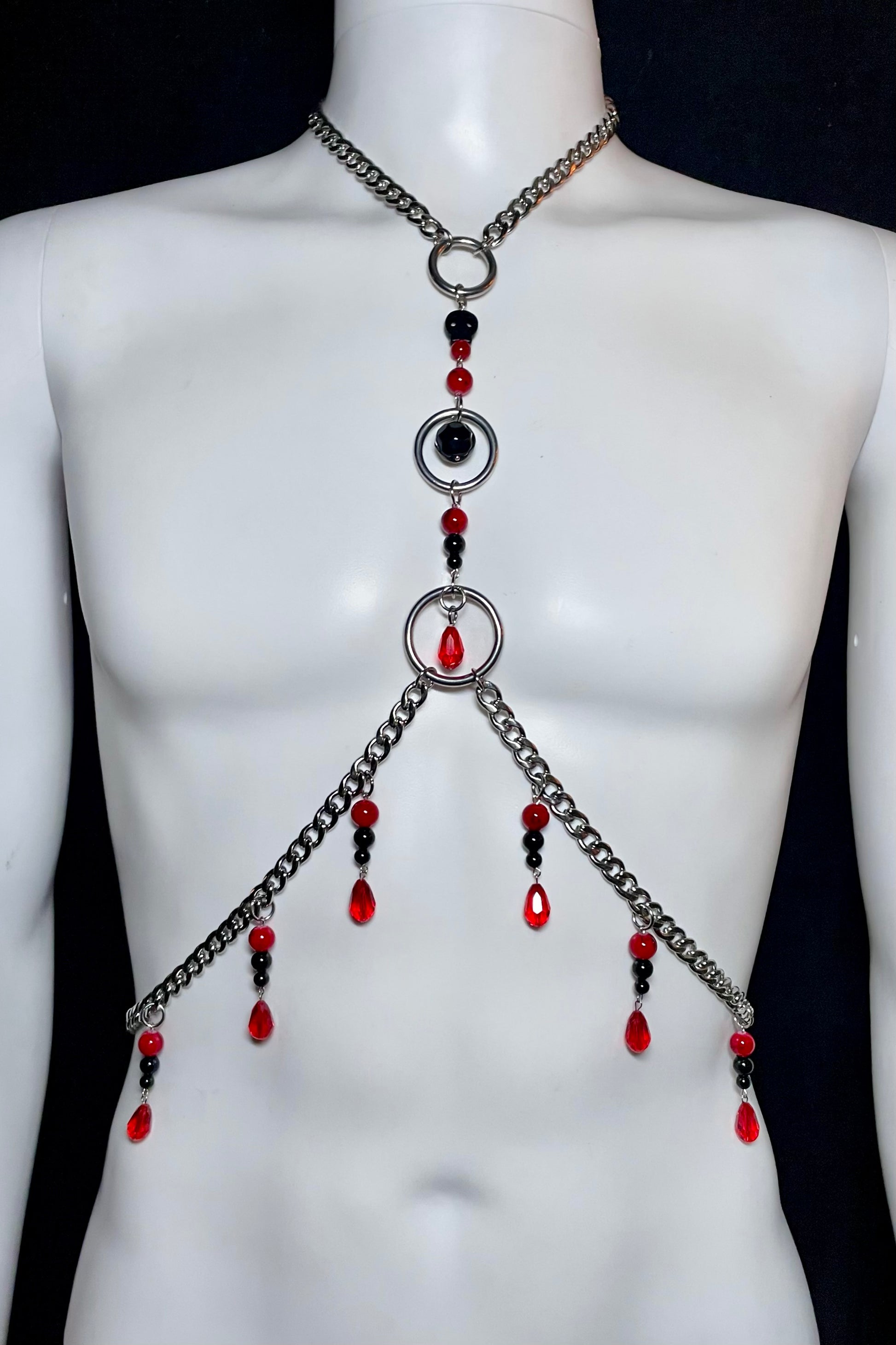 A stainless steel body harness featuring interlocking rings and adorned with red and black beads. Teardrop-shaped beads hang along the sides, creating a gothic, blood-inspired look.