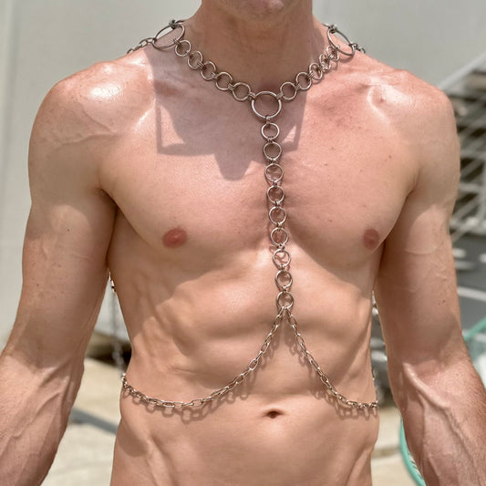 A shirtless model wearing a S
stainless steel chain body harness featuring a single line of circular links down the center of the chest. The design drapes around the shoulders and waist, creating a sleek and modern look perfect for festival wear or bold statement fashion.