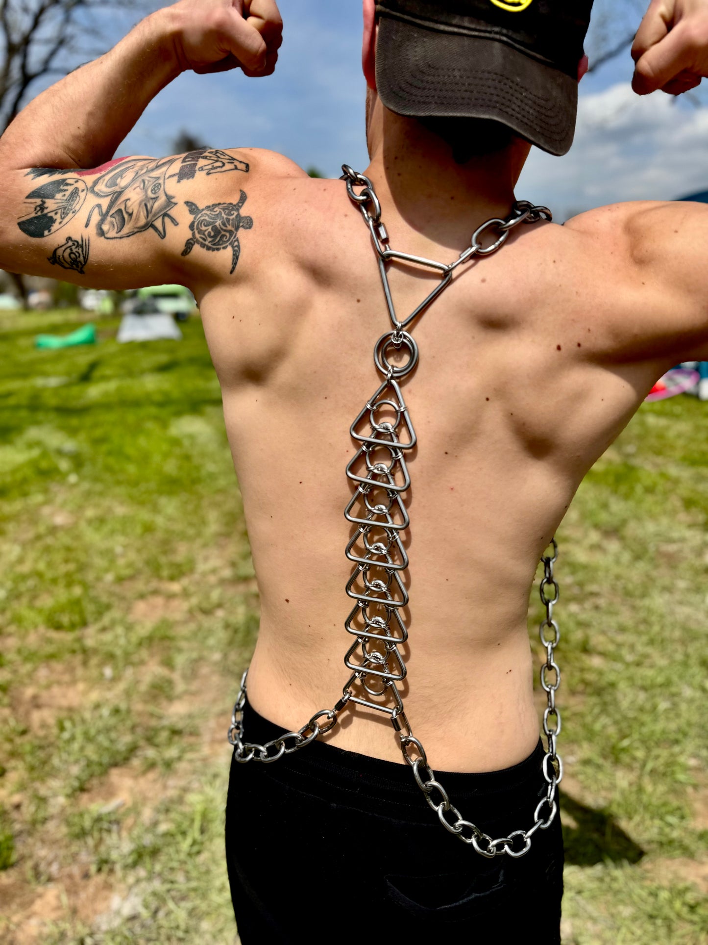 A shirtless model wearing a handcrafted stainless steel body harness featuring a bold geometric design of stacked triangles connected by chainmail links. The harness wraps around the shoulders and chest, paired with silver chains draped down the sides, creating a striking and edgy look.