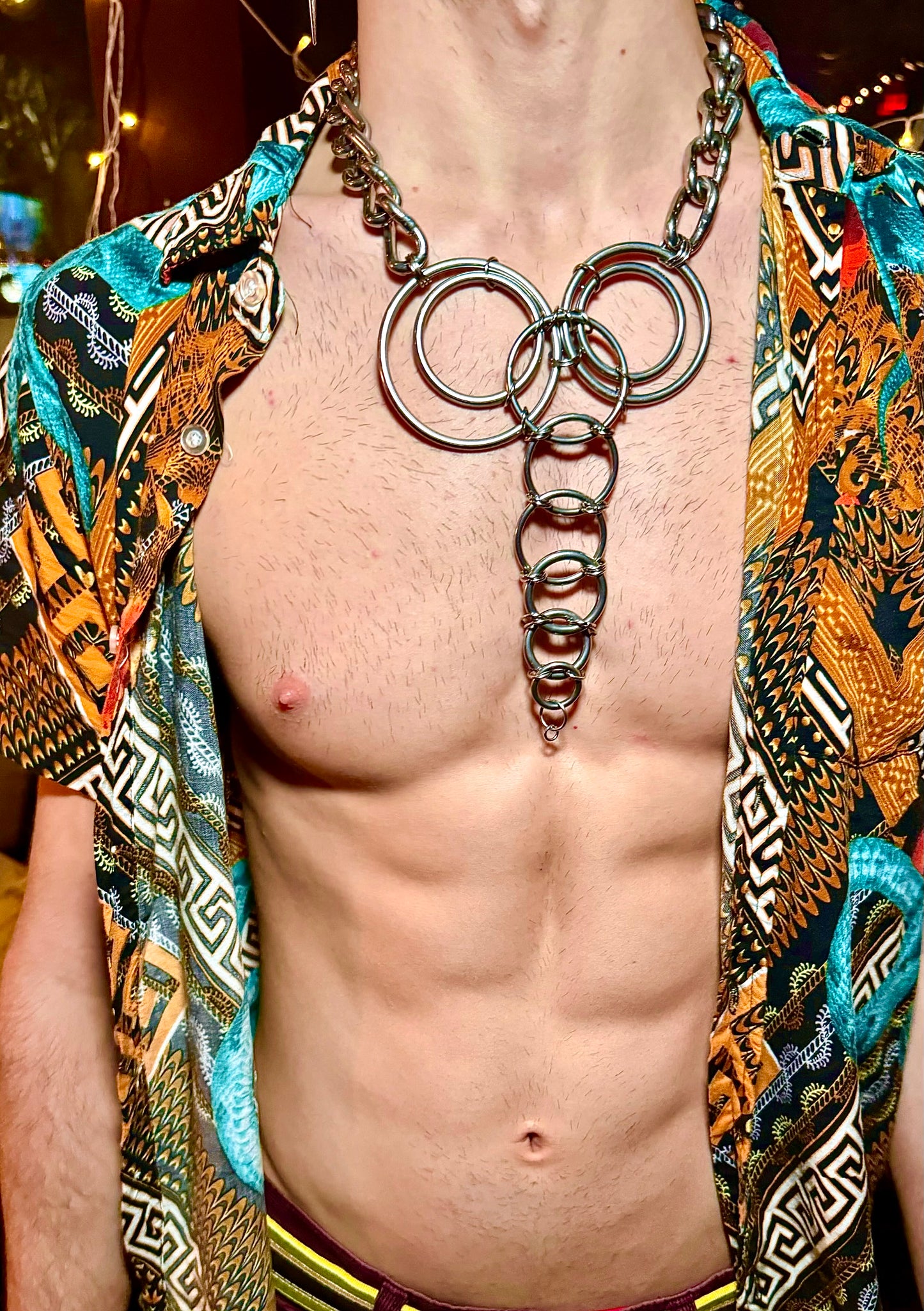 A model showcases a handcrafted stainless steel necklace with interlocking rings, forming a geometric trunk design across the chest. Paired with an open, colorful shirt, the necklace adds a bold, playful edge as a striking statement piece.