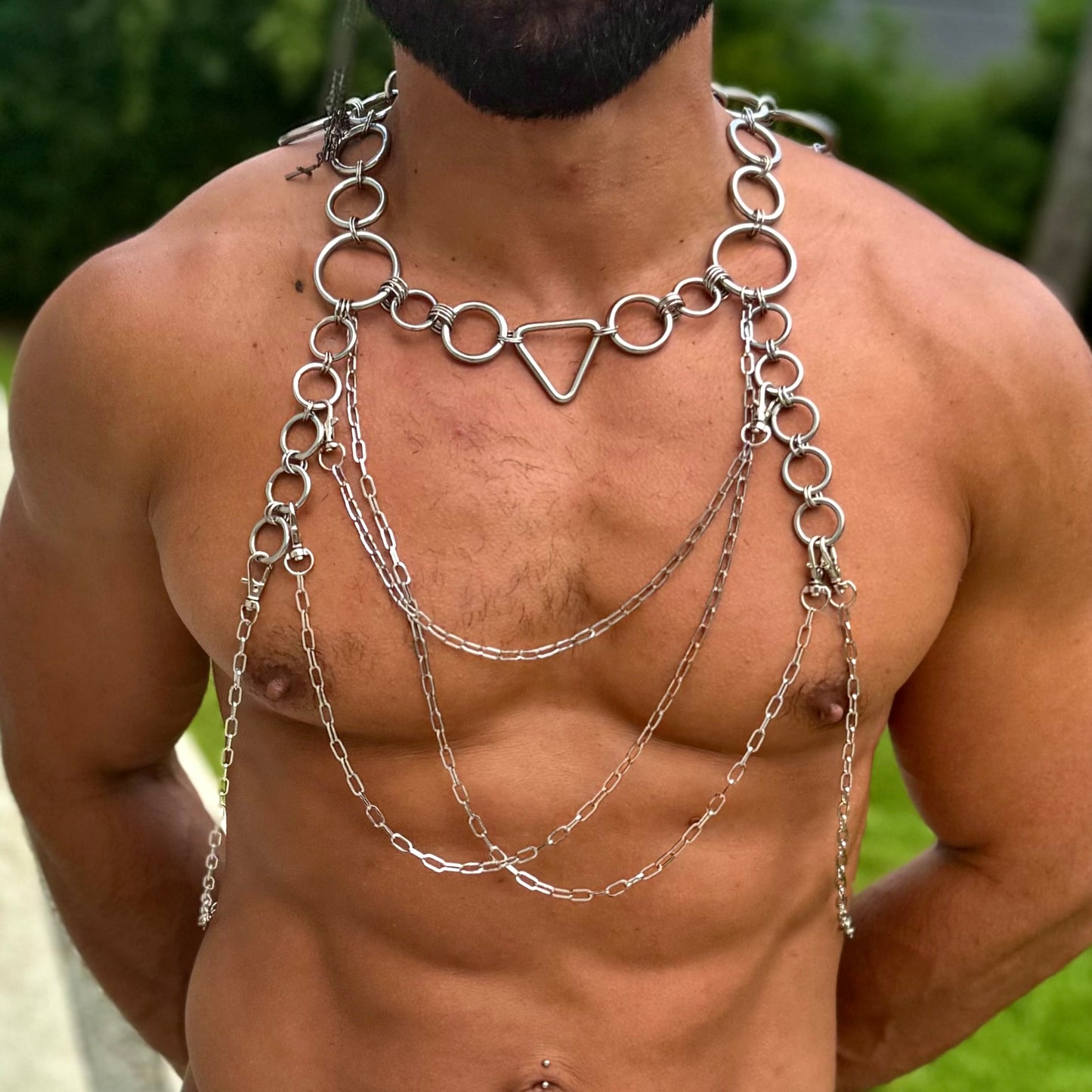 A person wearing an intricate body harness made of stainless steel rings and chains. The harness features a central triangle link at the neck, with multiple layers of chains draping across the chest and shoulders, creating a striking and artistic look. The design is bold, with circular rings connecting the chains and providing structure to the piece.