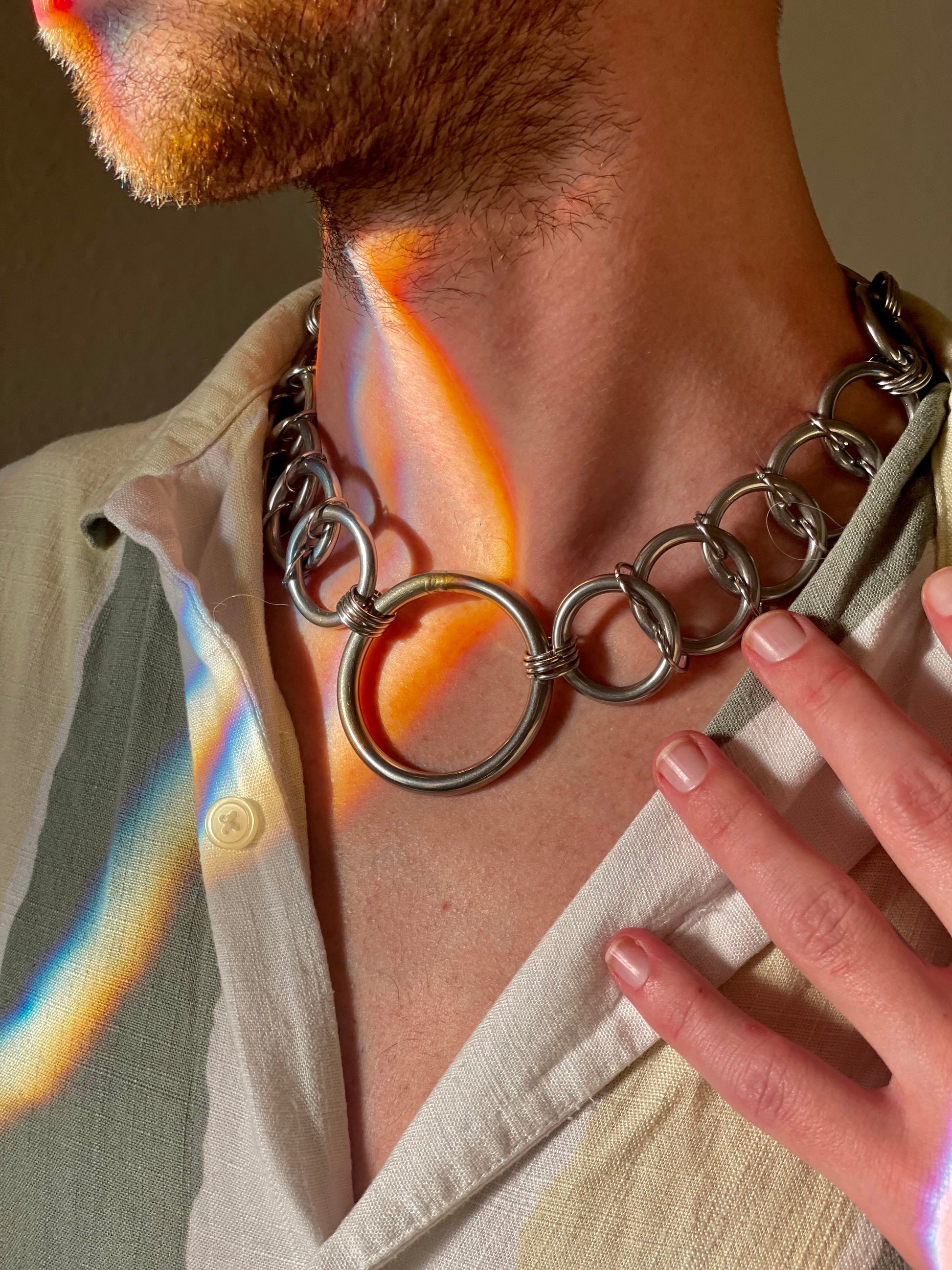 A close-up image of a person wearing a bold silver chain necklace featuring a large circular pendant in the center. The necklace shines in the warm, natural light, creating a striking contrast with the model’s light-colored shirt and the subtle rainbow hues on their skin.