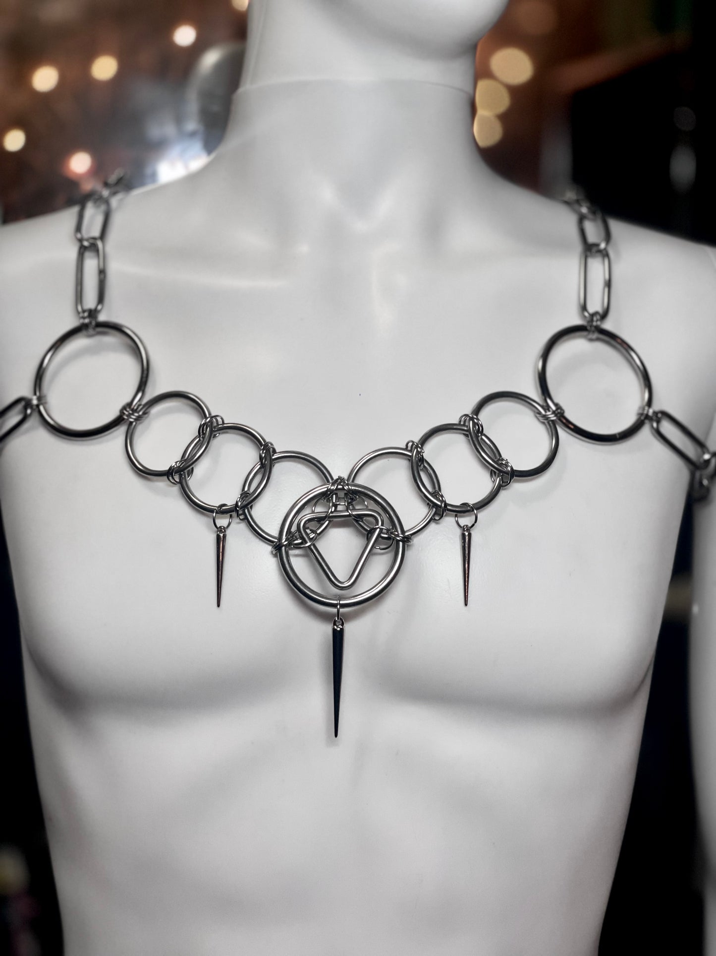 A mannequin wearing a bold stainless steel body chain featuring an array of circular rings across the chest. A triangle sits in the middle and draws the eye. The geometric design adds structure, while the layered chains create an industrial, artistic look. 