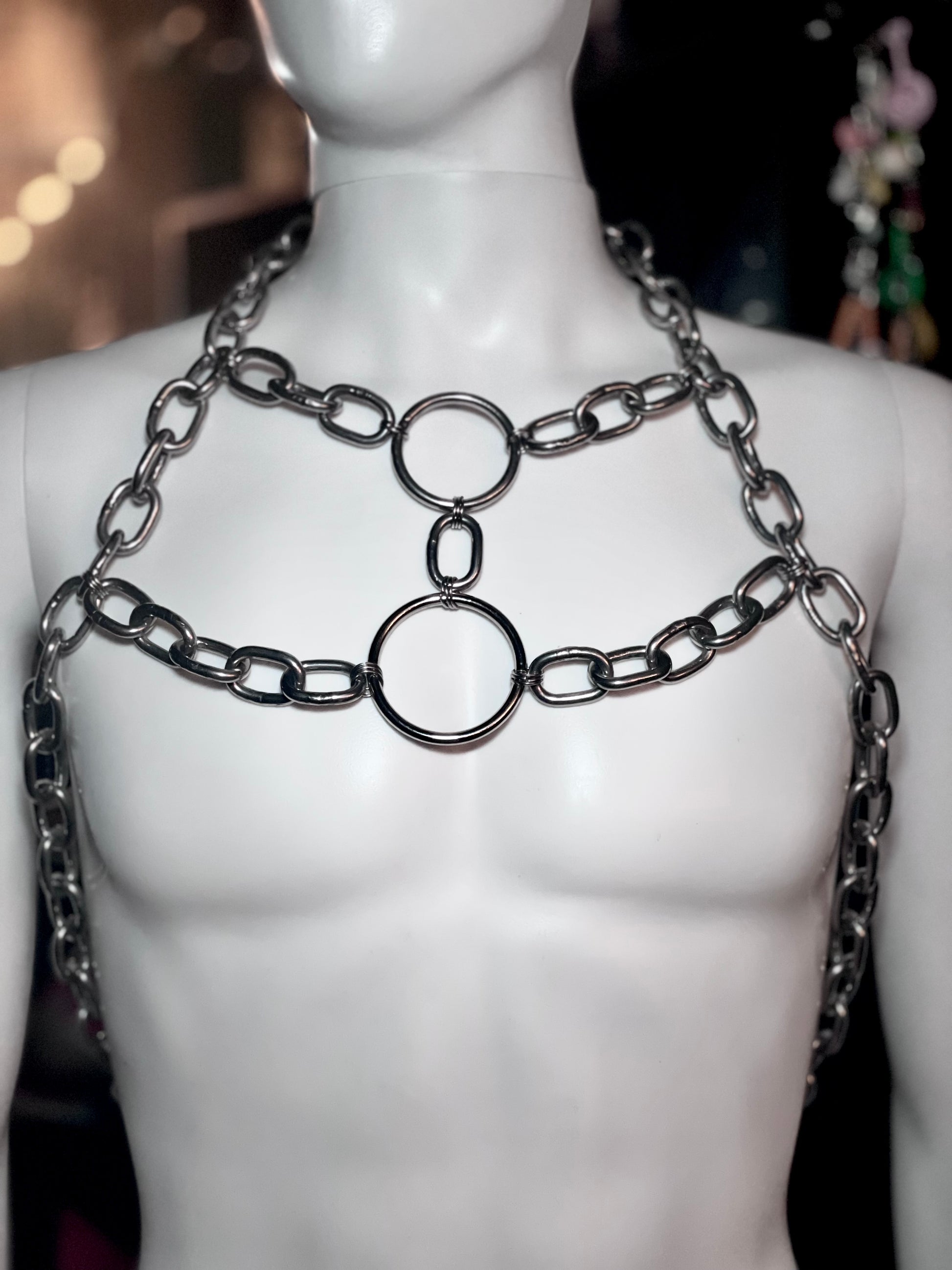 Close-up image of a bold, handmade stainless steel chain harness featuring interlocking circular rings and thick metal links. The geometric design drapes across the chest, showcasing an industrial yet artistic vibe perfect for statement fashion.